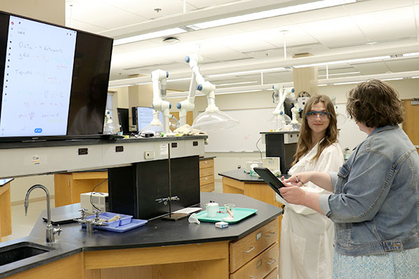 A student and a professor discuss a chemistry lab with an ipad and a large screen showing experiment notes.