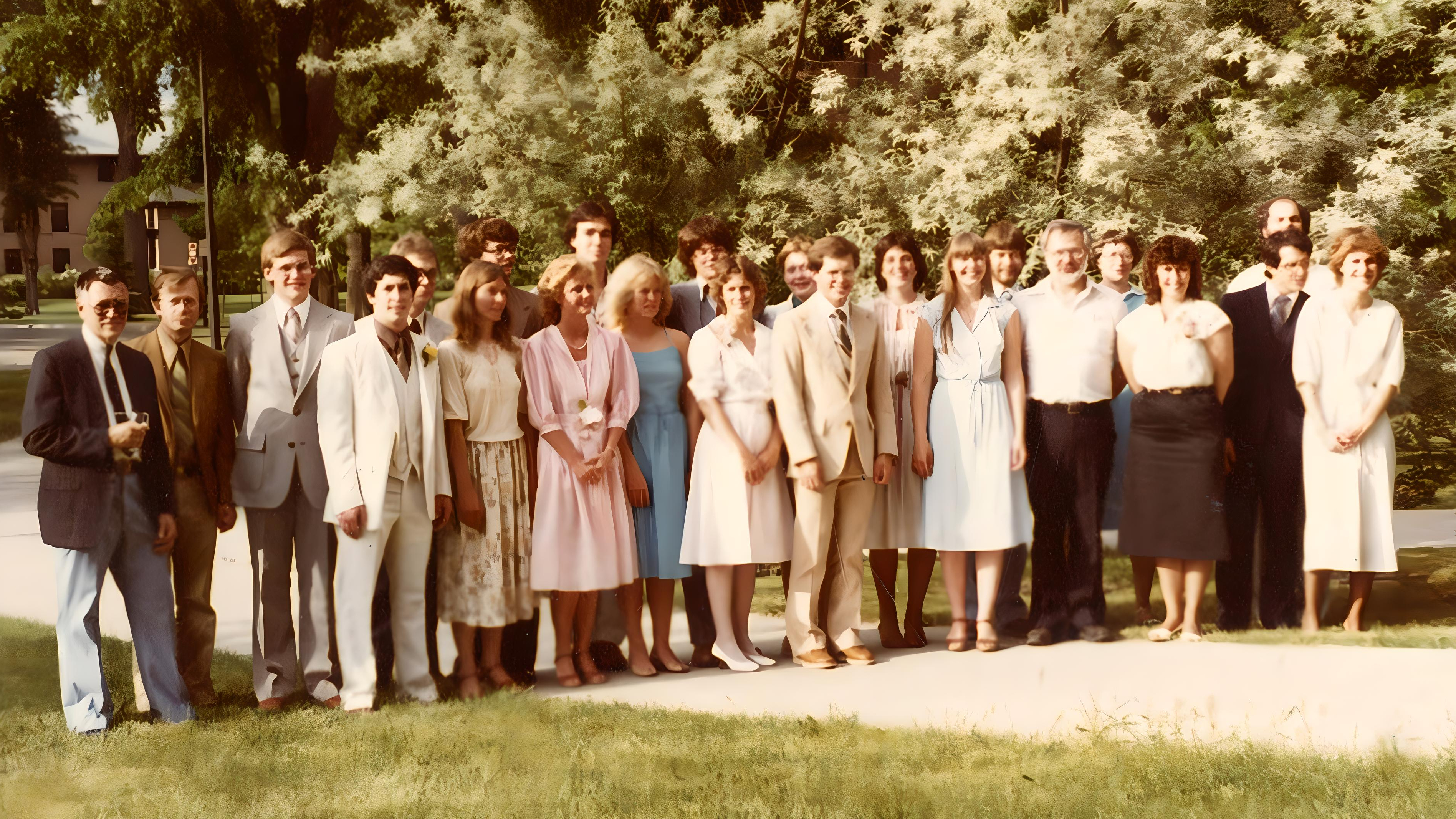 Biology majors class of 1983 with professors.  A group of nineteen people standing together in three rows on a grassy area, with trees in the background. Most are smiling and dressed in semi-formal attire.