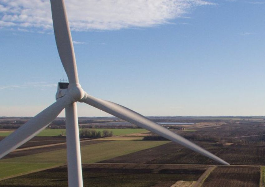 Close-up view of a large white wind turbine against a blue sky with light clouds, overlooking a vast landscape of agricultural fields.