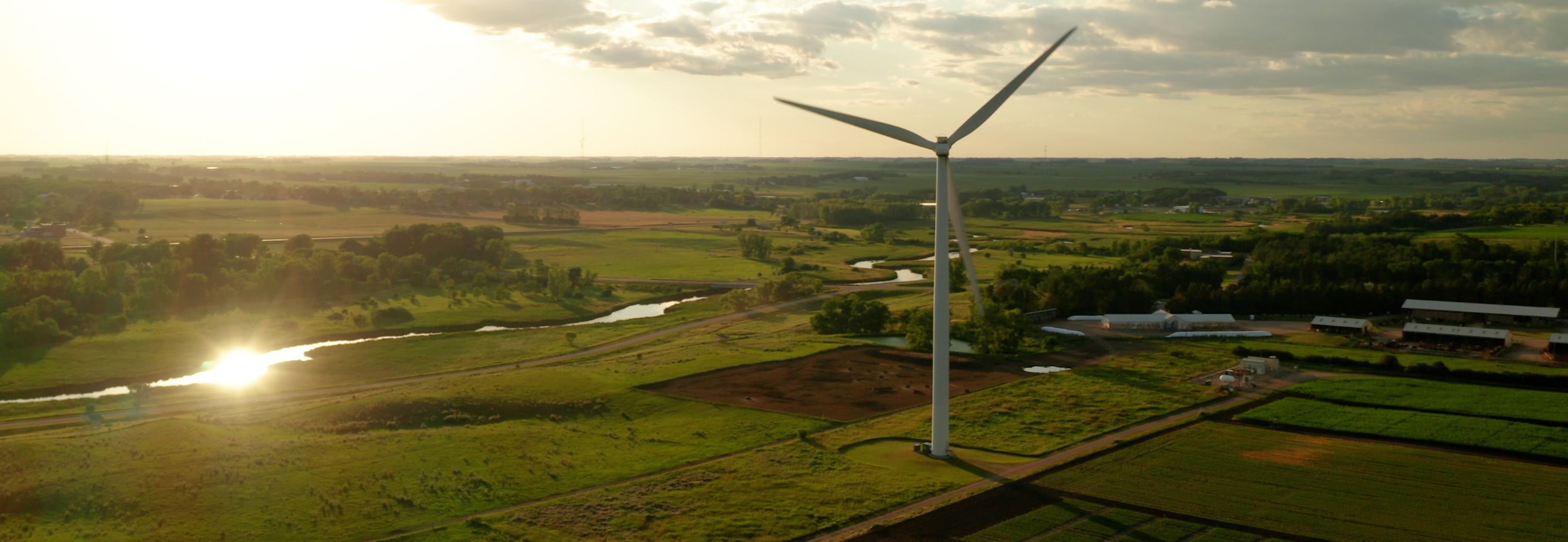 Aerial panoramic view of a large white wind turbine standing over green agricultural fields and a winding river reflecting the bright golden sunlight of a sunset.