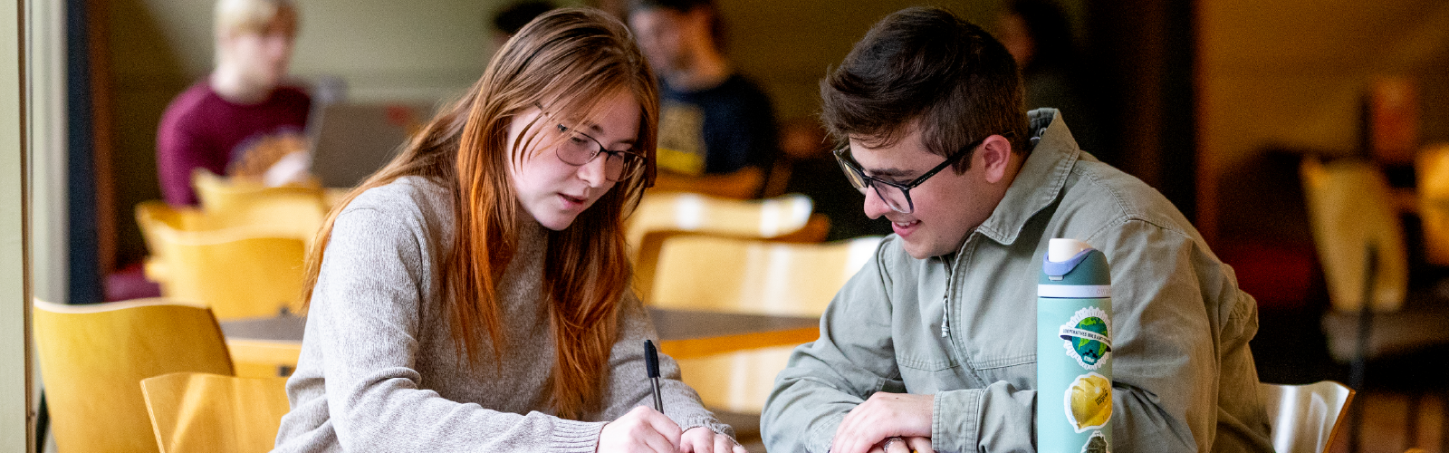 Two students sitting at a table together studying and taking notes in a campus dining area or student center.