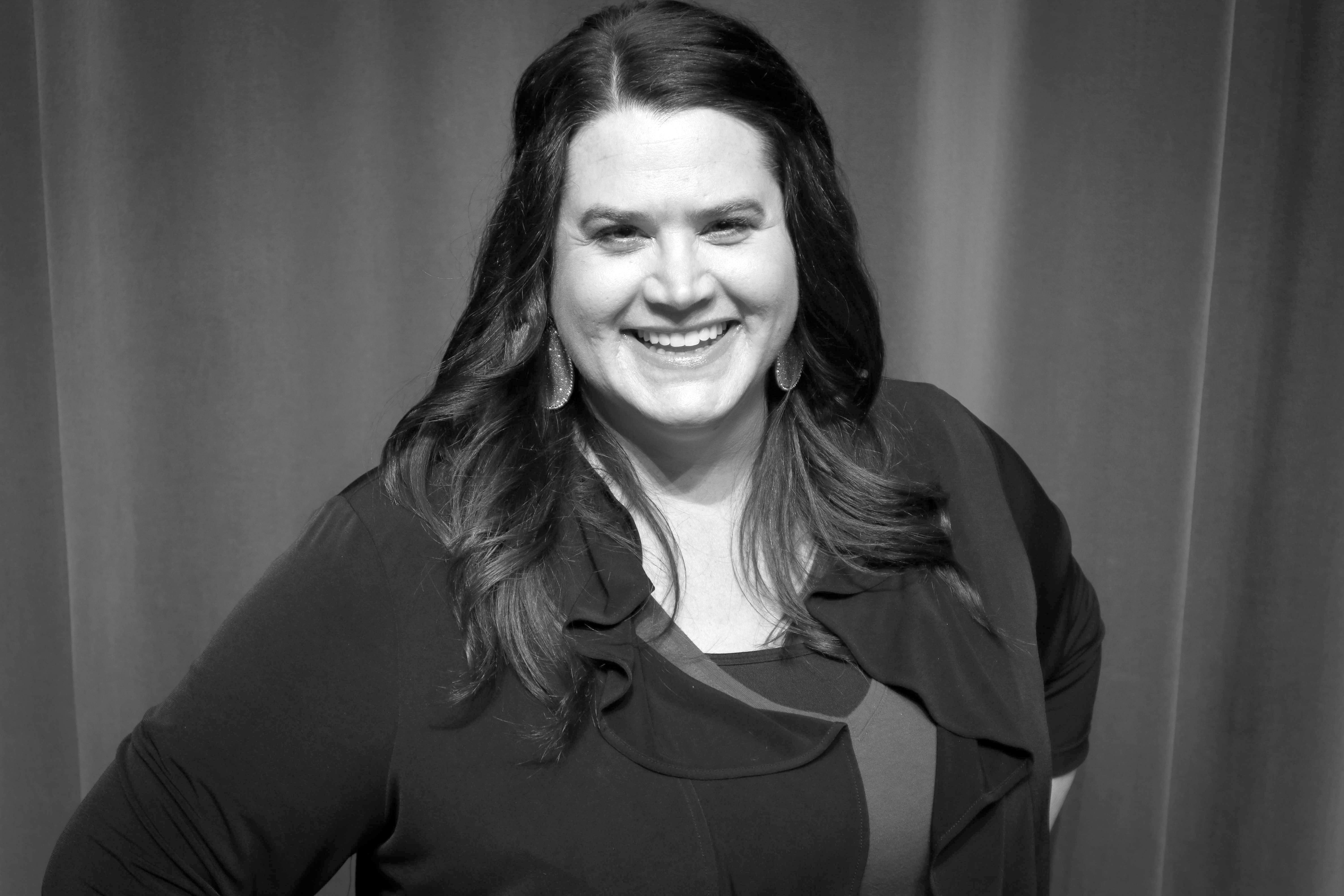 A black and white photo of a woman with long dark hair, hands on hips, smiling 