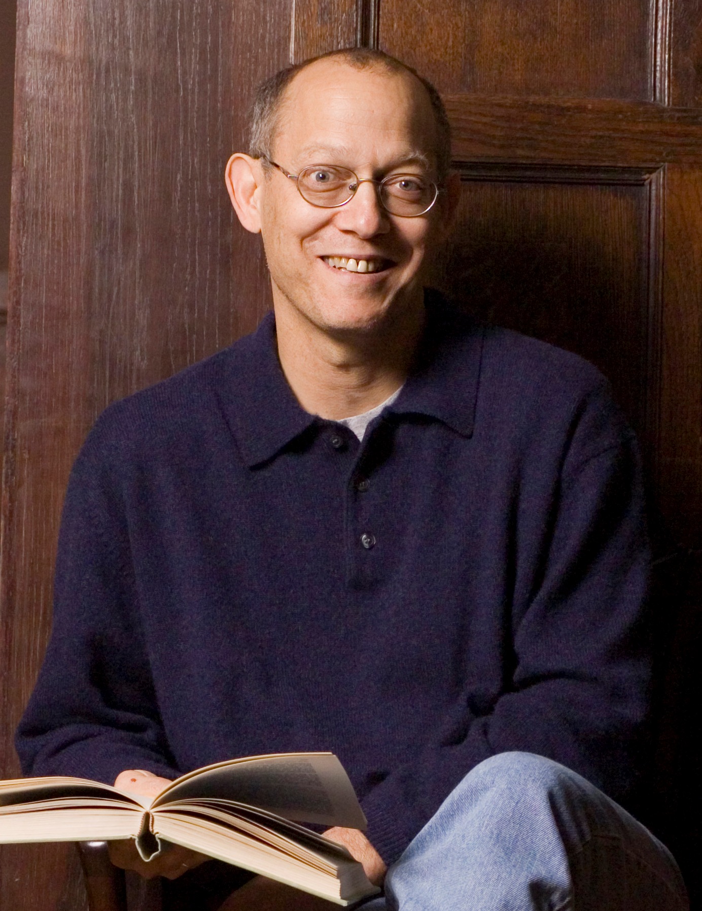 A white man wearing glasses and a purple shirt, holding an open book