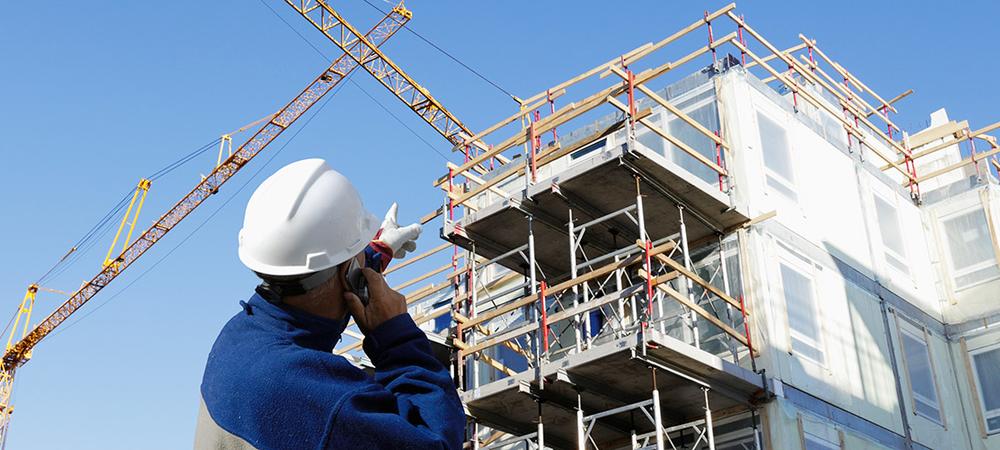 A man wearing a hardhat gestures and speaks on a cell phone in front of a building under construction.