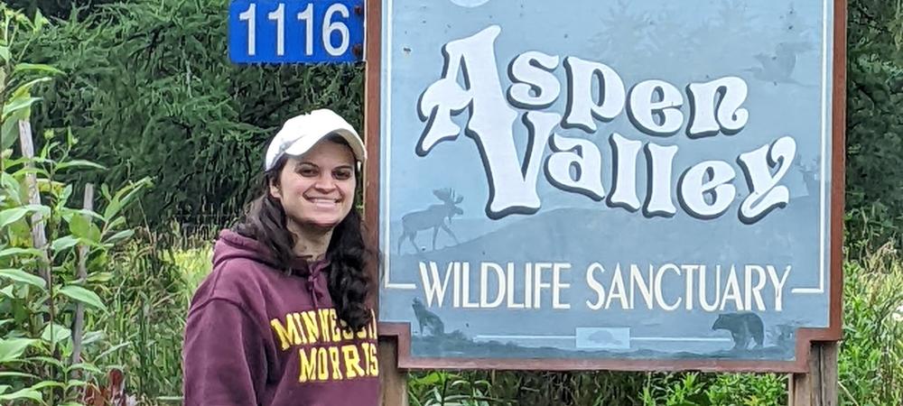 A man wearing a grey pants, maroon sweatshirt and a white baseball cap standing beside a sign that reads, Aspen Valley Wildlife Sanctuary