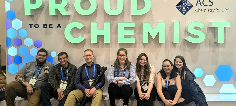 Seven people sitting in front of a wall with the worlds, Proud To Be a Chemist