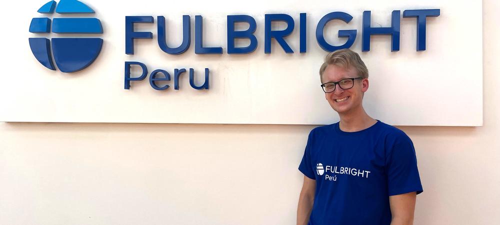 A blonde man with glasses wearing a blue t-shirt and black pants, standing in front of a sign that reads, Fulbright Peru