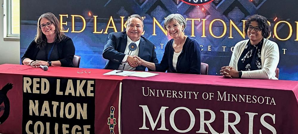 Four people sitting at a table.  The two in the center are shaking hands.  The table coverings read "Red Lake Nation College" and University of Minnesota Morris.  