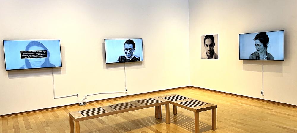 An series of television monitors and photographs on the wall, showing people, with wooden benches placed near them. 