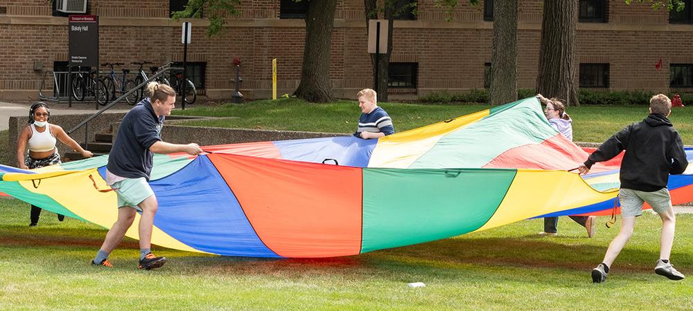 A group of students plays a game with a large, colorful parachute on the campus mall.
