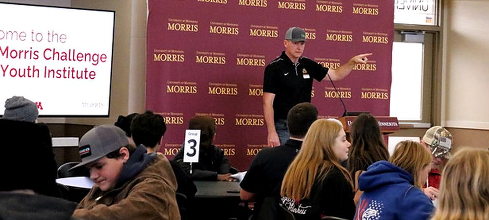 A white man in a cap and black polo shirt standing in front of a group of high school students. 