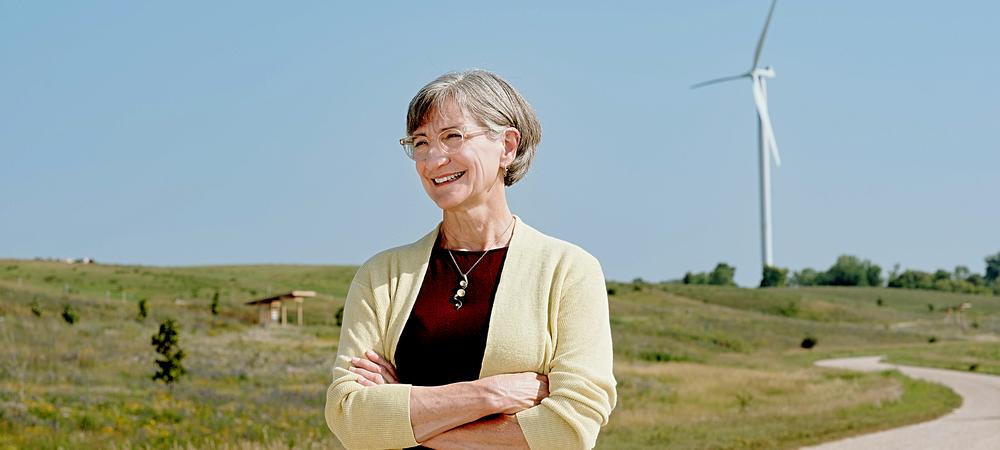 A white woman with gray hair wearing a maroon shirt and pale yellow cardigan, standing on a gravel road with a prairie landscape in the background.  In the distance, there is a white wind turbine. 