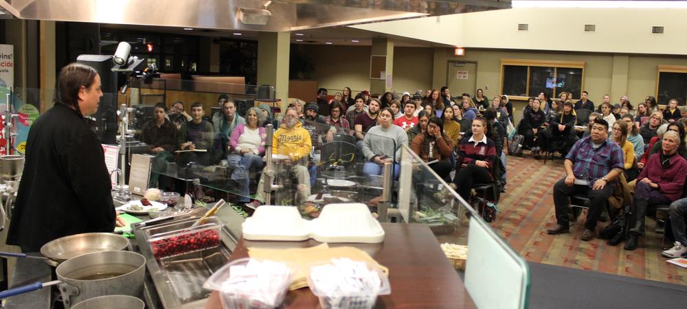 A Native American man, with long braids, wearing a black chef's jacket, standing at a cook's station, with an audience watching him. 