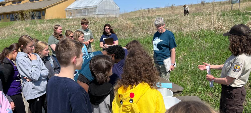 A group of high school students in a field, listening to a staff member from the U.S. Fish and Wildlife Service.  There is a building in the background.  