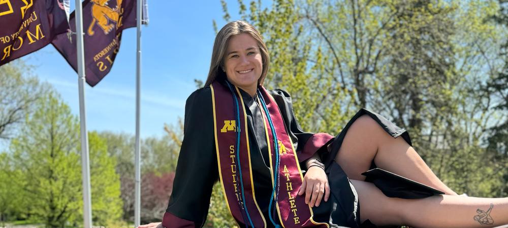 A person in graduation attire sits on a brick wall. The person is wearing a black graduation gown and cap, and has a sash with "MORRIS" printed on it. Several maroon and gold university flags flutter in the background. The setting is outdoors on a sunny day.