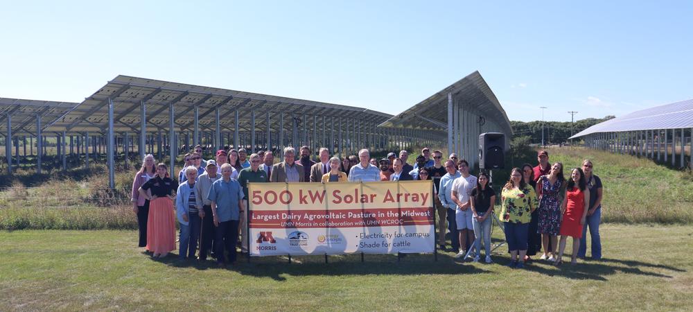 Group of people holding a banner celebrating the inauguration of a 500 kW solar array, the largest dairy agrivoltaic pasture in the Midwest, with solar panels and a clear sky in the background.