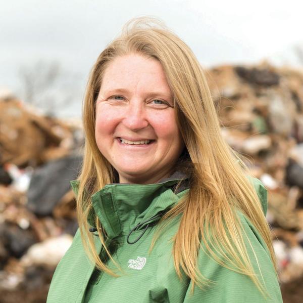 A smiling woman with long blonde hair, wearing a green rain coat, standing in front of a pile of trash