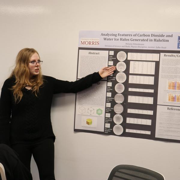 A woman standing by a scientific poster, pointing to its contents, with another woman sitting