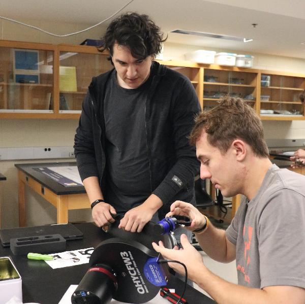 Two college students are working on electronic equipment in a laboratory setting. One person is using a soldering iron on a device, while the other observes closely. The table is scattered with various tools and components. 