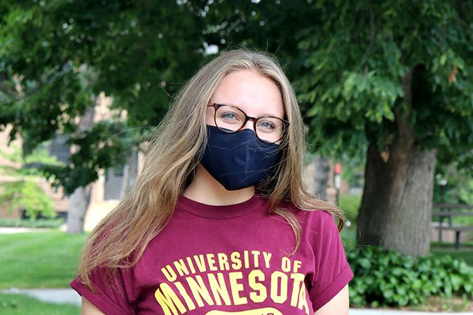 a student wearing a mask stands near the campus mall and smiles into lens