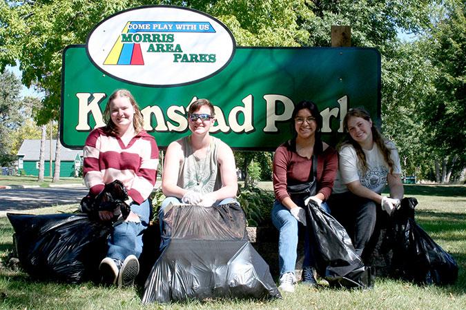Four students pose with bags of trash picked up during a park cleanup at a Morris Area Parks sign for Kjenstad Park.