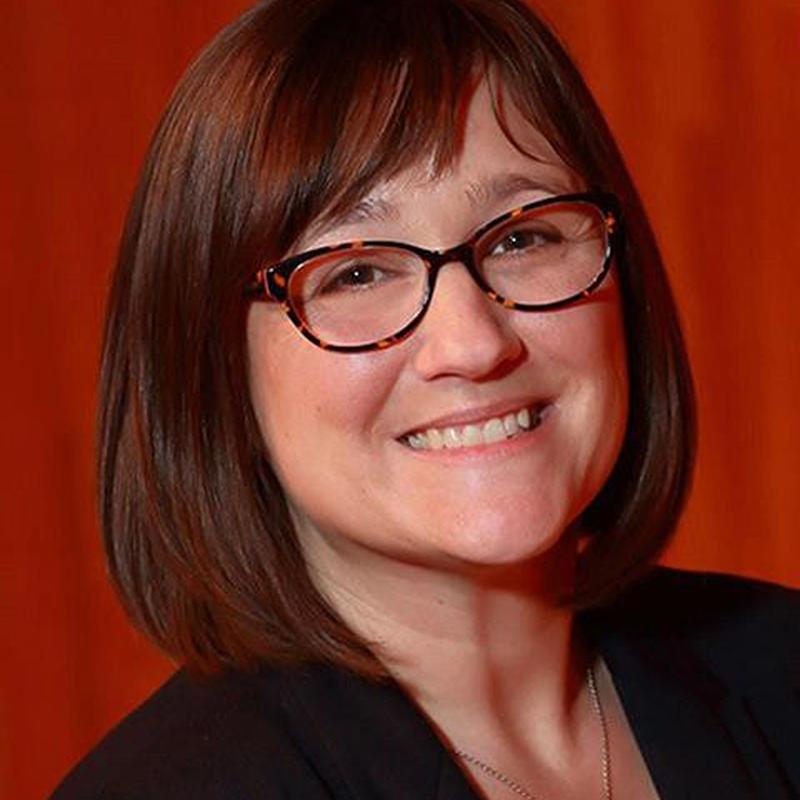 A white woman with brown hair, wearing glasses and a black blazer against a red background