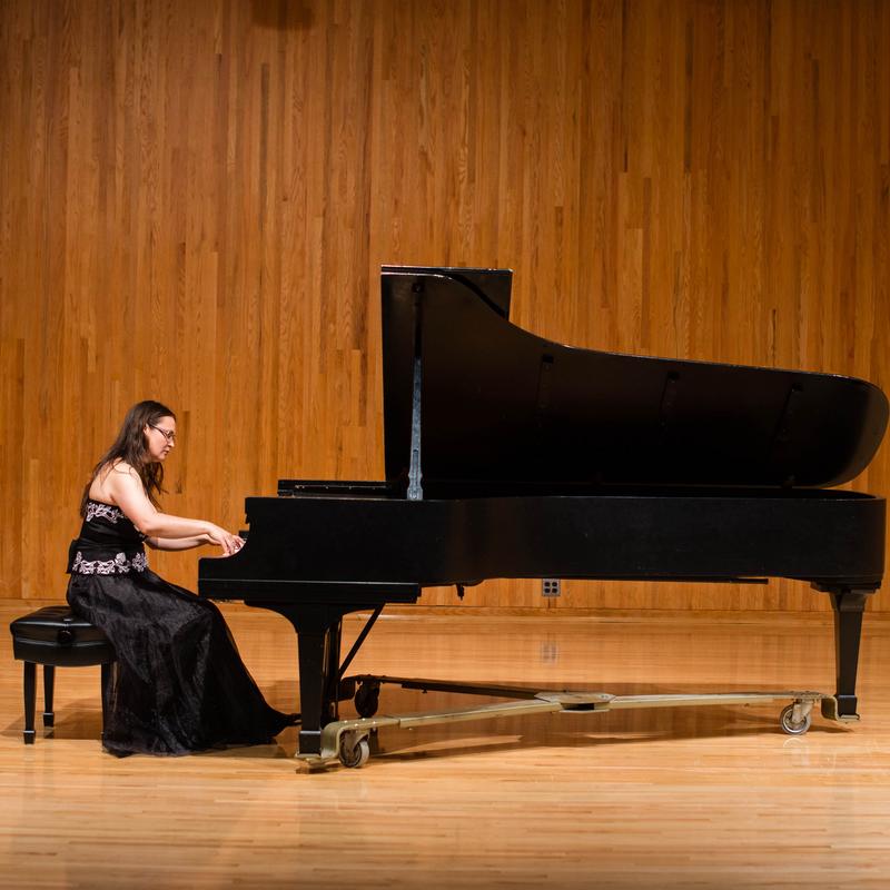 A woman with long dark hair and glasses, wearing a sleeveless black ball gown, playing a grand piano.
