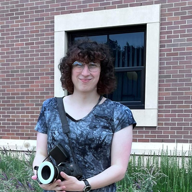 A woman with dark curly hair and glasses wearing a blue and black swirled t-shirt and holding a camera.  She is standing in front of flowers by a brick building.