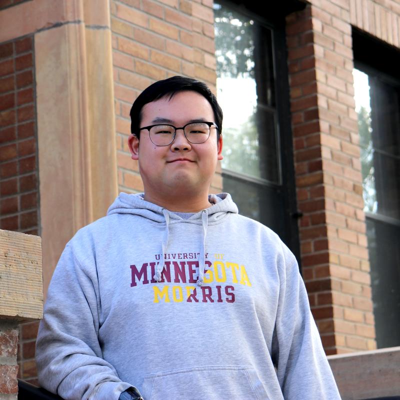 A young Chinese man standing outside of a brick building.  He has short black hair, and black-rimmed glasses and is wearing a grey sweatshirt with the words, University of Minnesota Morris on it. 