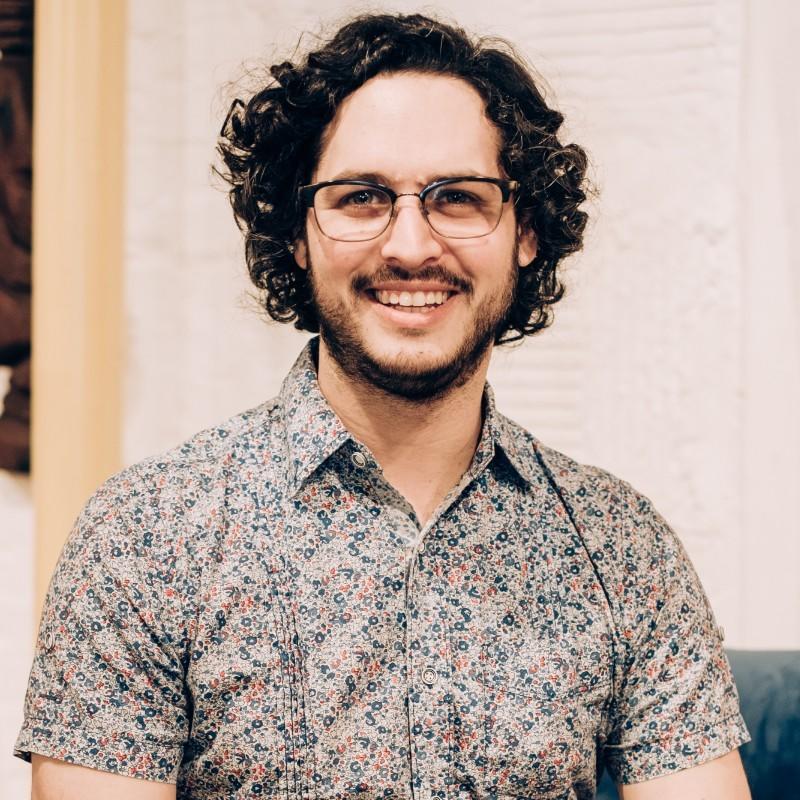 A man with short curly hair, and short beard, wearing glasses and a patterned shirt