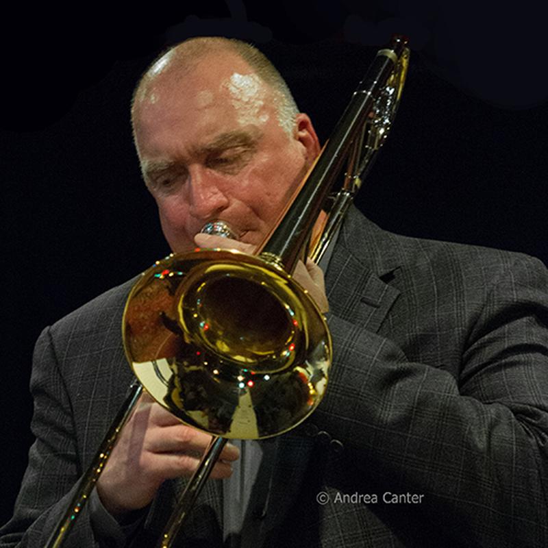 A white man with balding hair, wearing a grey suit jacket, playing a trombone