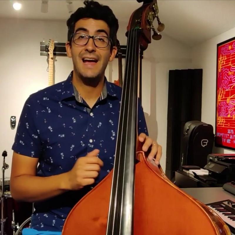 A smiling man with short dark hair and glasses, wearing a blue patterned shirt and holding a standing bass