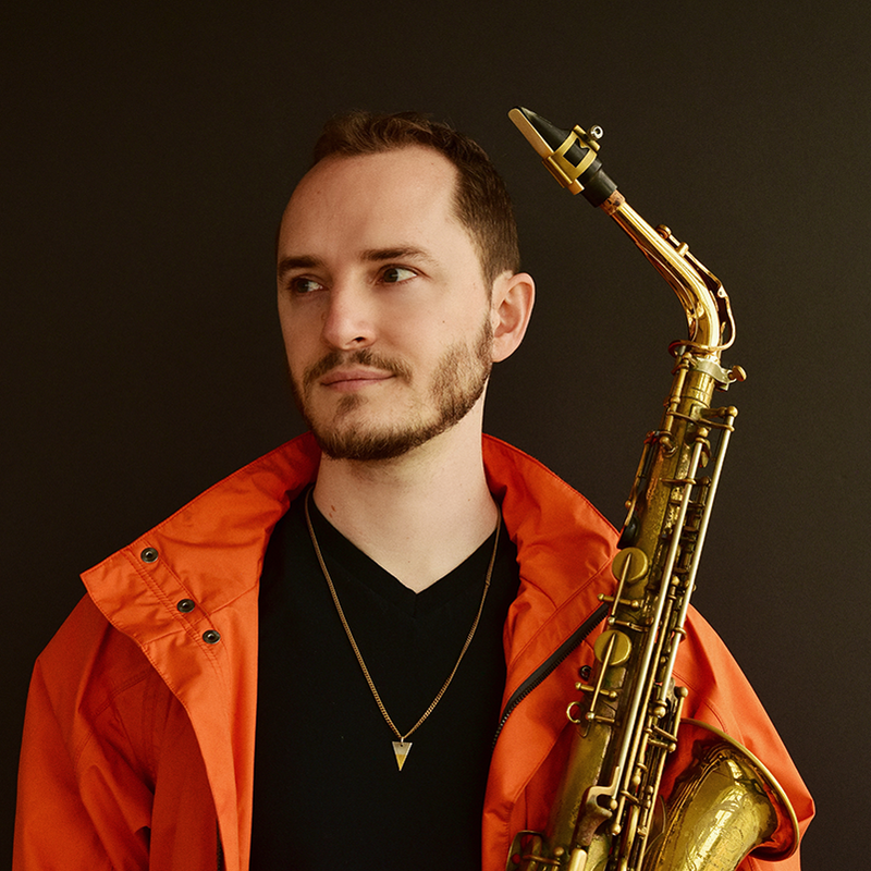 A man with short hair and a tightly trimmed beard, wearing an orange jacket and black t-shirt.  He is holding an alto saxophone