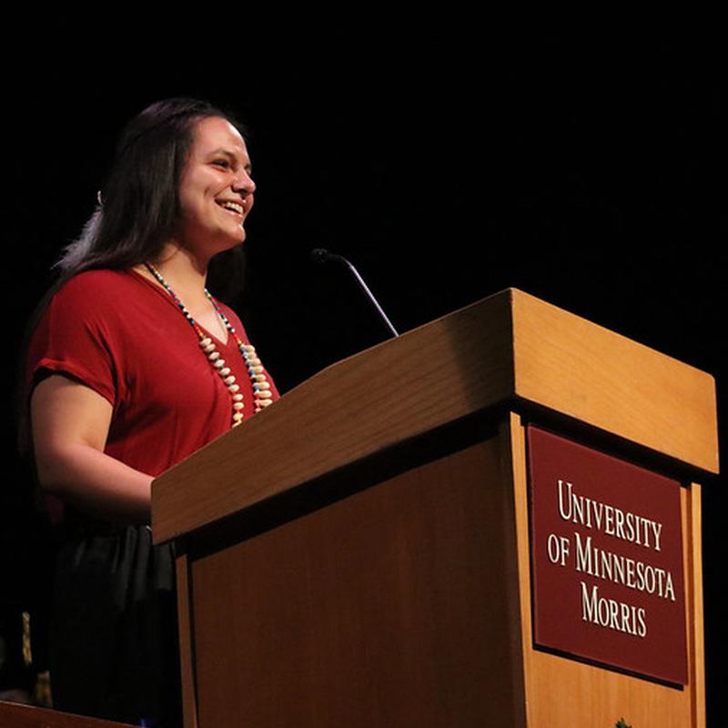 An Indigenous woman with long black hair, wearing a red shirt, standing at a podium 