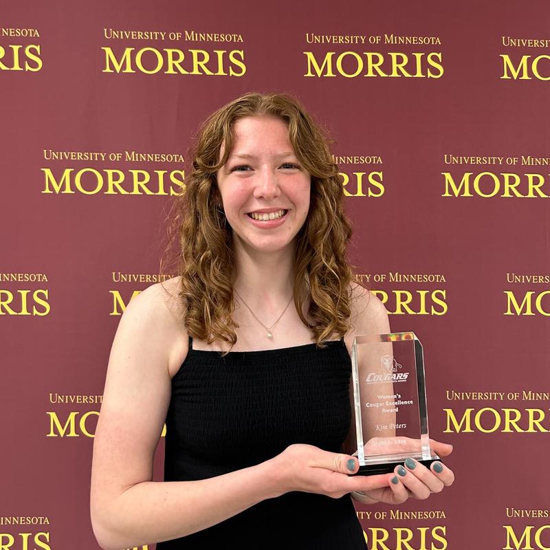 A young woman with long brown hair, wearing a black, spaghetti strap dress and holding an award. She is standing in front of a backdrop that repeatedly says University of Minnesota Morris