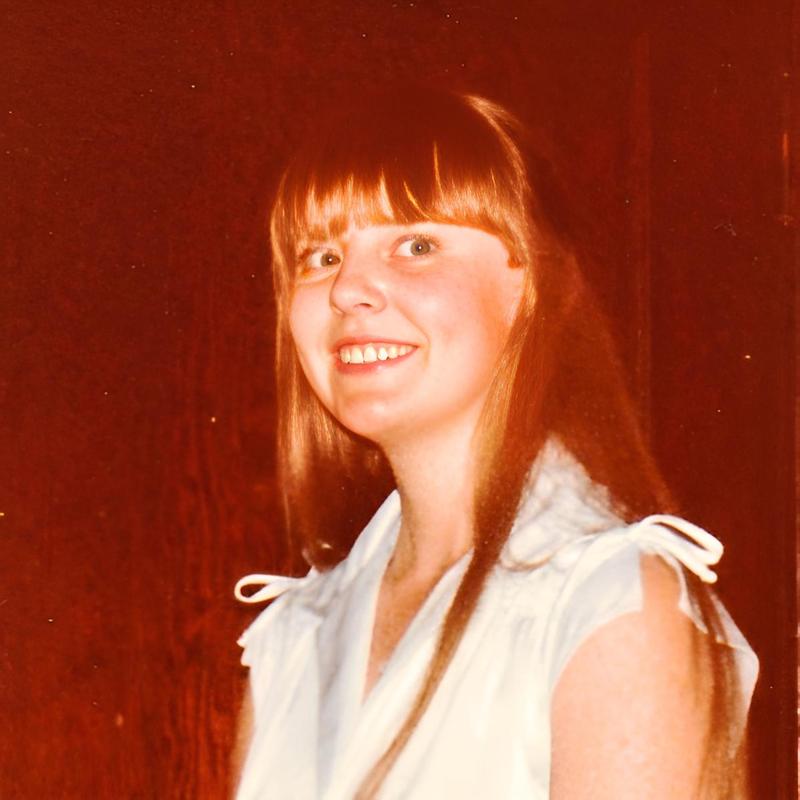 A vintage photo of Jane Johnson, a woman with long, straight hair and bangs, smiling broadly. She wears a light-colored sleeveless top with tie details on the shoulders. The background is a plain, dark color which highlights the subject.