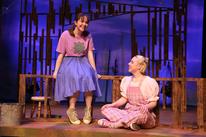 Two young women on a stage, one wearing a t-shirt and denim skirt and sitting on a bench, the other in pink overalls and sitting on the floor. 