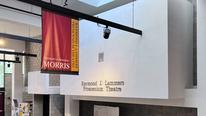 A white wall with the words, Raymond J. Lammers Proscenium Theatre writte on it, with a maroon and gold banner hanging in front. 