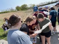 A group of five high school students huddled around a piece of paper.  They are outside in a grassy field.  There is another group of students behind them. 