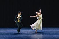 Two ballet dancers perform on stage, one in a flowing beige dress and the other in a black suit with a gold vest, expressing dramatic emotion through their graceful movements.