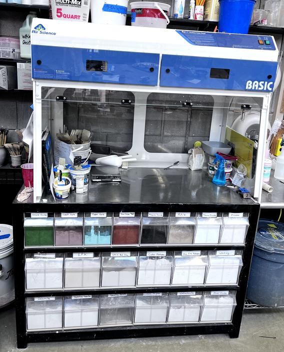 A glazing station situated above a storage unit filled with labeled bins of various colored resins. The station is in a workspace, surrounded by various supplies and tools.