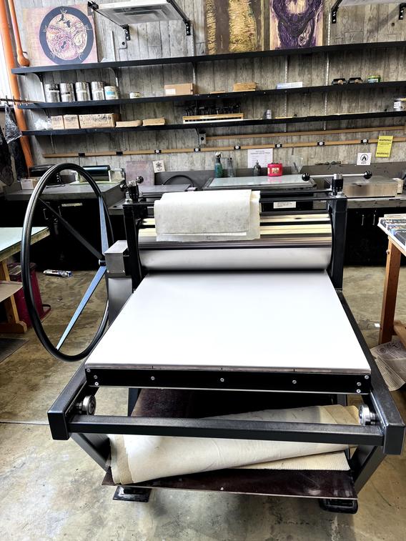 A printmaking press with a large roller, a flat bed, and a crank handle, located in an artistic studio with various supplies and artwork visible in the background.