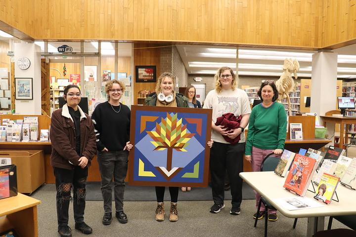 Four college students and the library director displaying a barn quilt