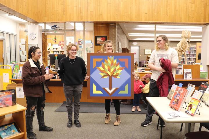 Four college students and a toddler displaying a barn quilt at the Morris Public Library 