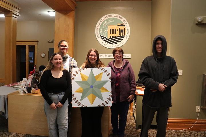 Four college students and their professor displaying a barn quilt at the Stevens County History Museum  