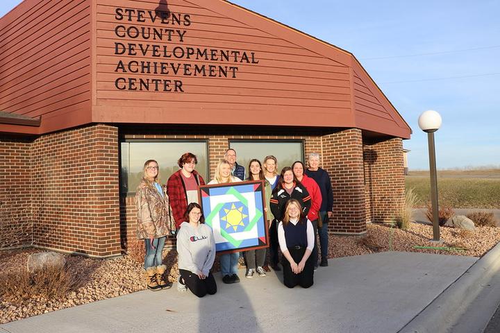 Five college students and five staff from the Stevens County DAC in front of their building holding a barn quilt 