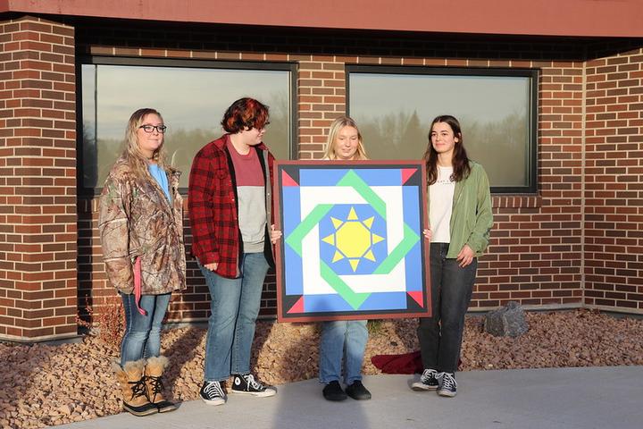 Four college students holding a barn quilt in front of a brick building. 
