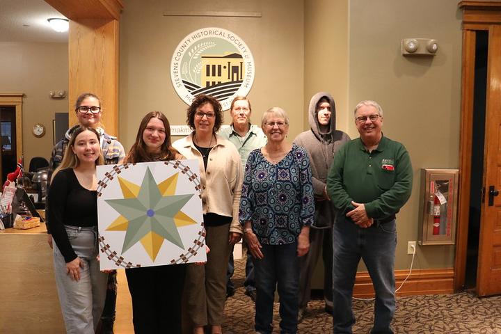 Four college students with the director, a staff member and two members of the board of directors displaying a barn quilt at the Stevens County History Museum