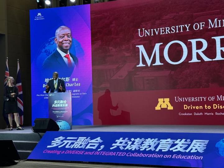 A black man standing at a podium in front of a large photo of himself. There are words in Chinese on the front of the stage. 