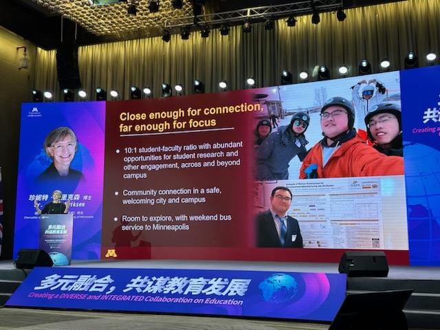 A woman at a podium in front of a very large screen with photos and text.  There is a message in Chinese in front of the stage